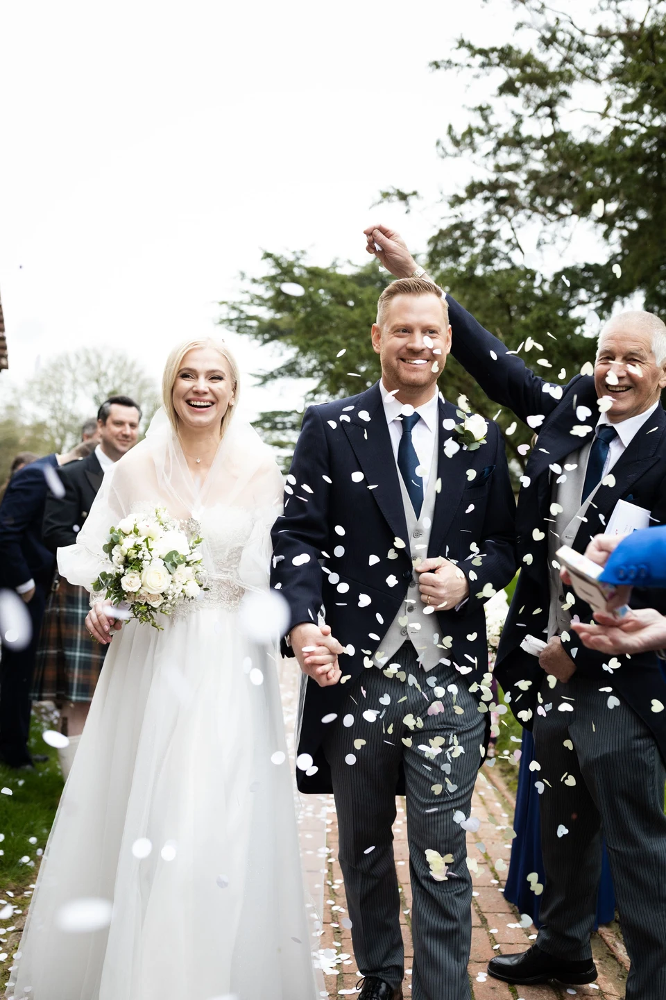 Vira and David smiling as guests throw confetti outside St Mary’s Church in Newick — Sussex